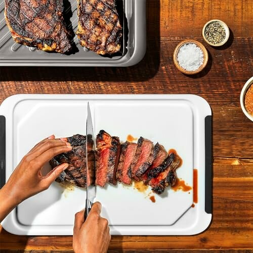 Person slicing a grilled steak on a white cutting board with seasonings nearby.