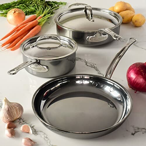 Stainless steel cookware set with vegetables on marble countertop.